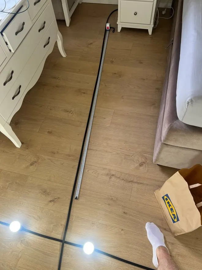 A tape measure along the floor between a white chest of drawers and the bed, next to the black cable.