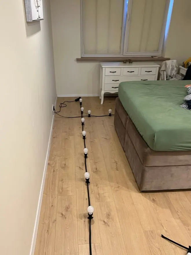 The string light on the floor along the bed, with plain bulbs in the holders and the plug in the wall socket.