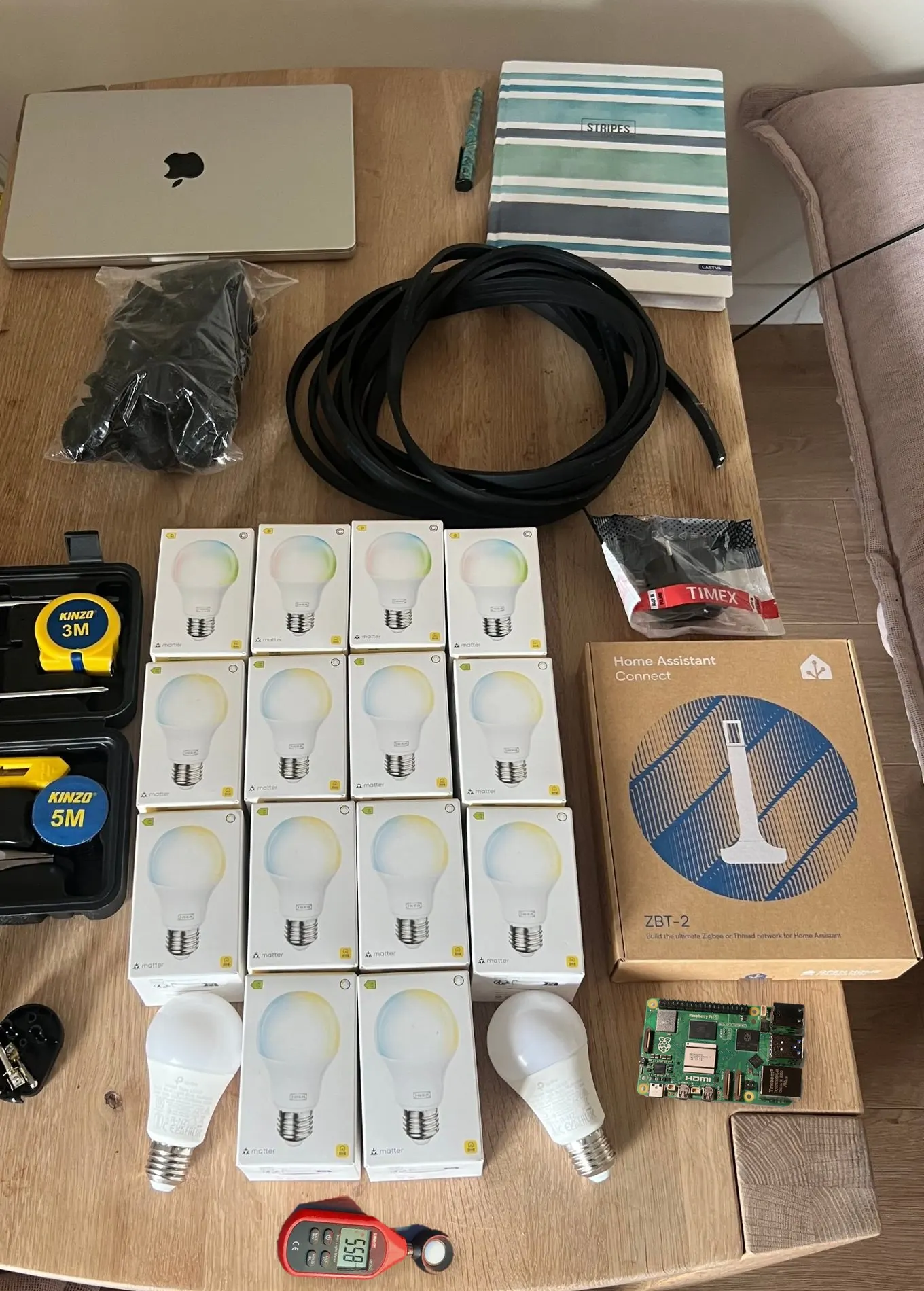Everything for the build on a table: a coil of flat black cable, a bag of lamp holders, boxes of IKEA KAJPLATS bulbs, a plug in its bag, a Home Assistant Connect ZBT-2 box, a Raspberry Pi 5, a UNI-T UT383 lux meter and a tool kit, next to a laptop and a notebook.
