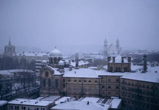 Snowy rooftops and church towers of Vilnius under a flat, hazy sky.